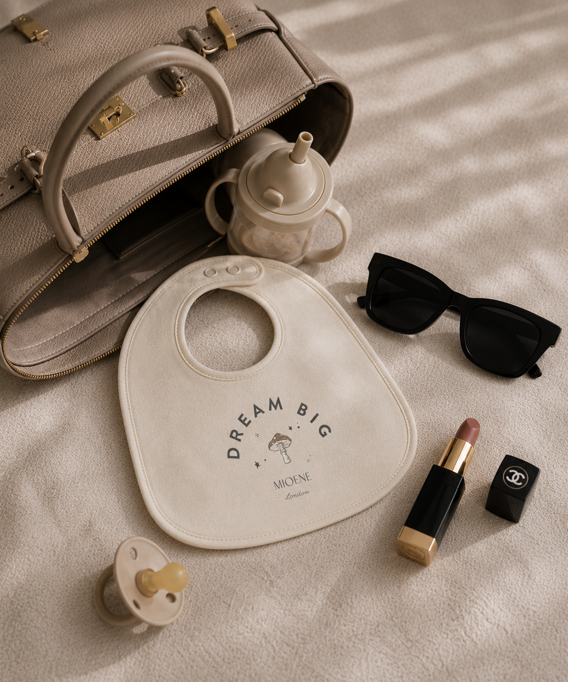Baby bib with 'Dream Big' text, baby bottle, sunglasses, lip balm, and lipstick on a textured surface.